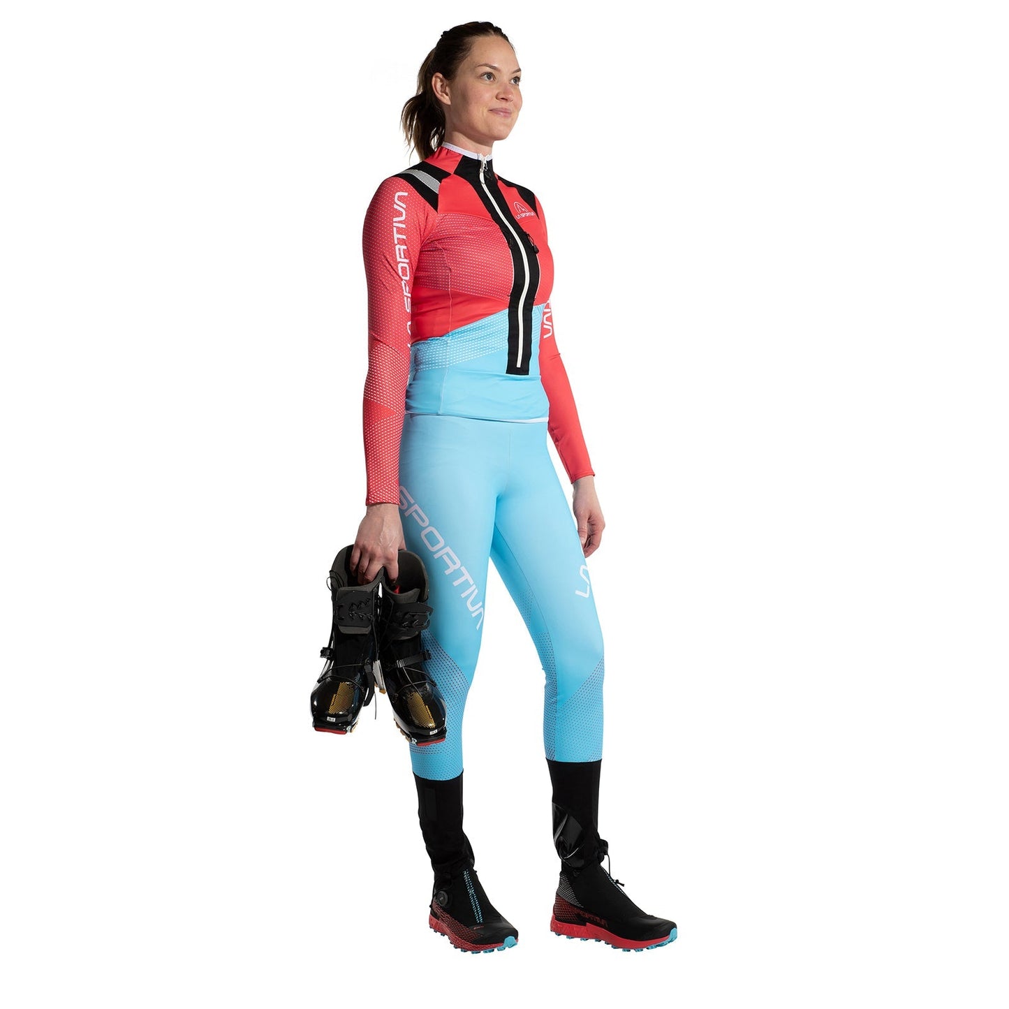 A woman wears the La Sportiva Stratos VI Racing Jacket Women's, made from stretch fabric in red and light blue. She stands holding black ski boots in her right hand and wears black and red athletic shoes, ready for fast slope training.