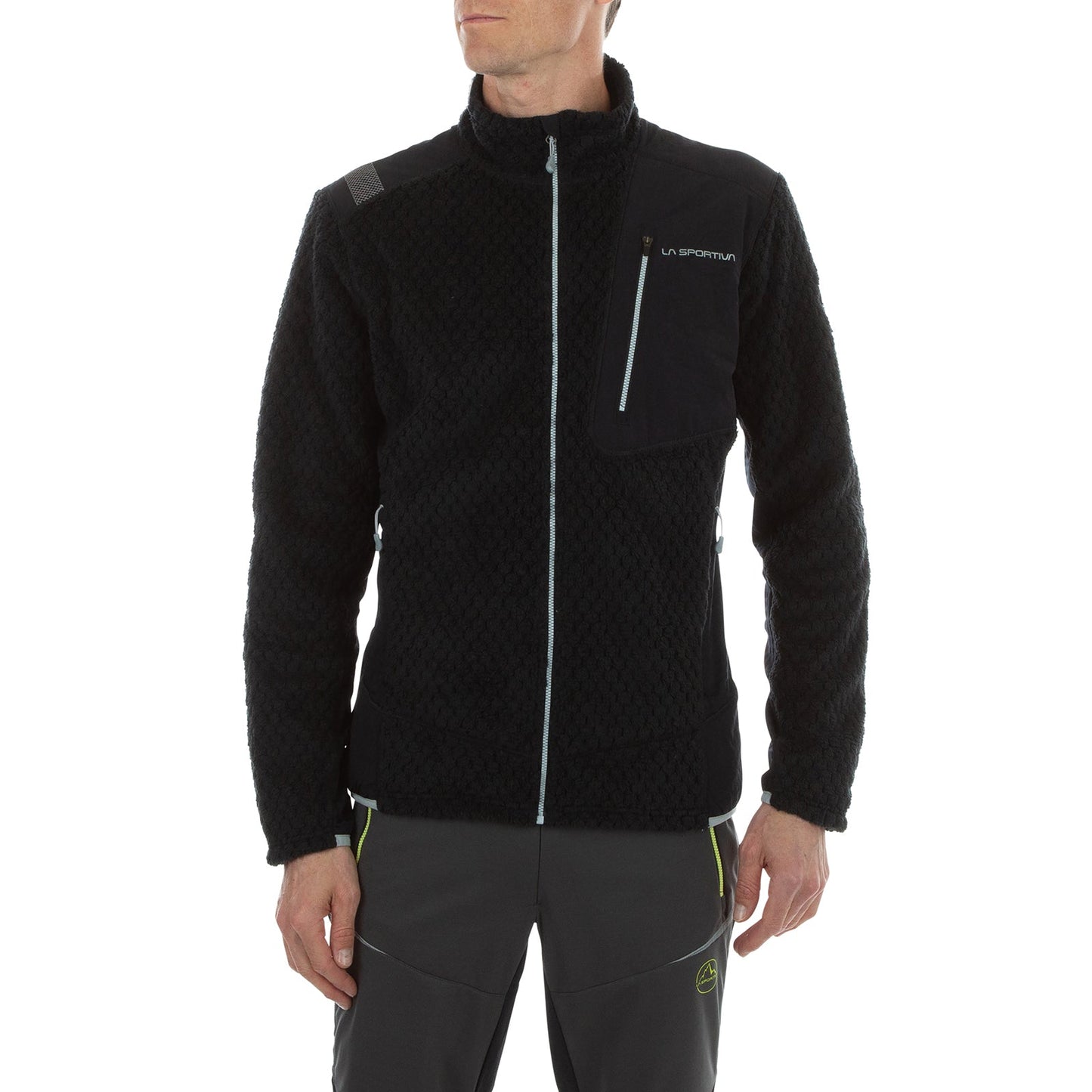 A man wearing the La Sportiva Bristen Thermal Jacket Men’s in black with a chest pocket and dark gray pants stands with arms relaxed at his sides, facing slightly left against a white background.