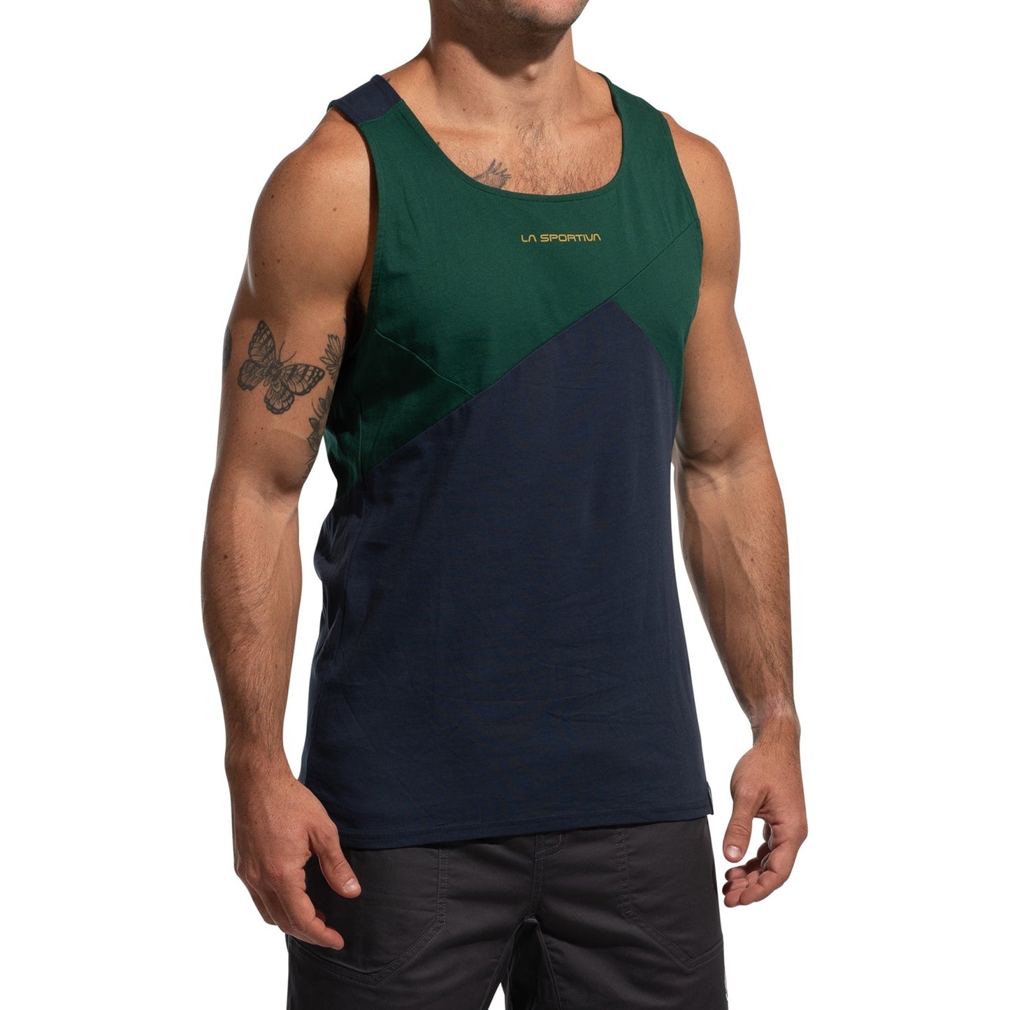 A man wearing the La Sportiva Dude Tank Men's (2025) climbing top in green and navy, featuring "La Sportiva" on the chest, with dark shorts and visible left-arm tattoos, stands against a plain white background.