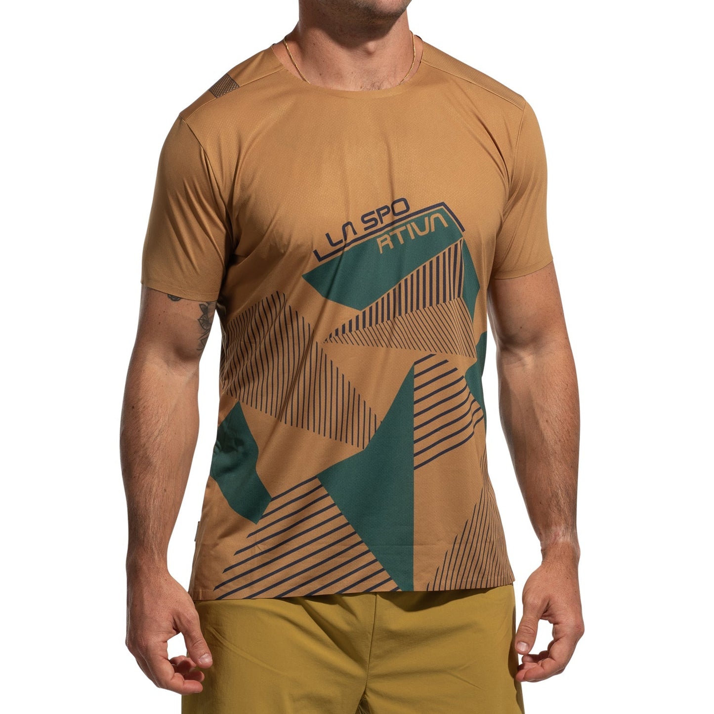 A man stands with arms relaxed at his sides, wearing the La Sportiva Comp T-Shirt Men's (2025), a lightweight tan short-sleeve top with green geometric patterns and "LA SPORTIVA" on the front, paired with tan shorts.