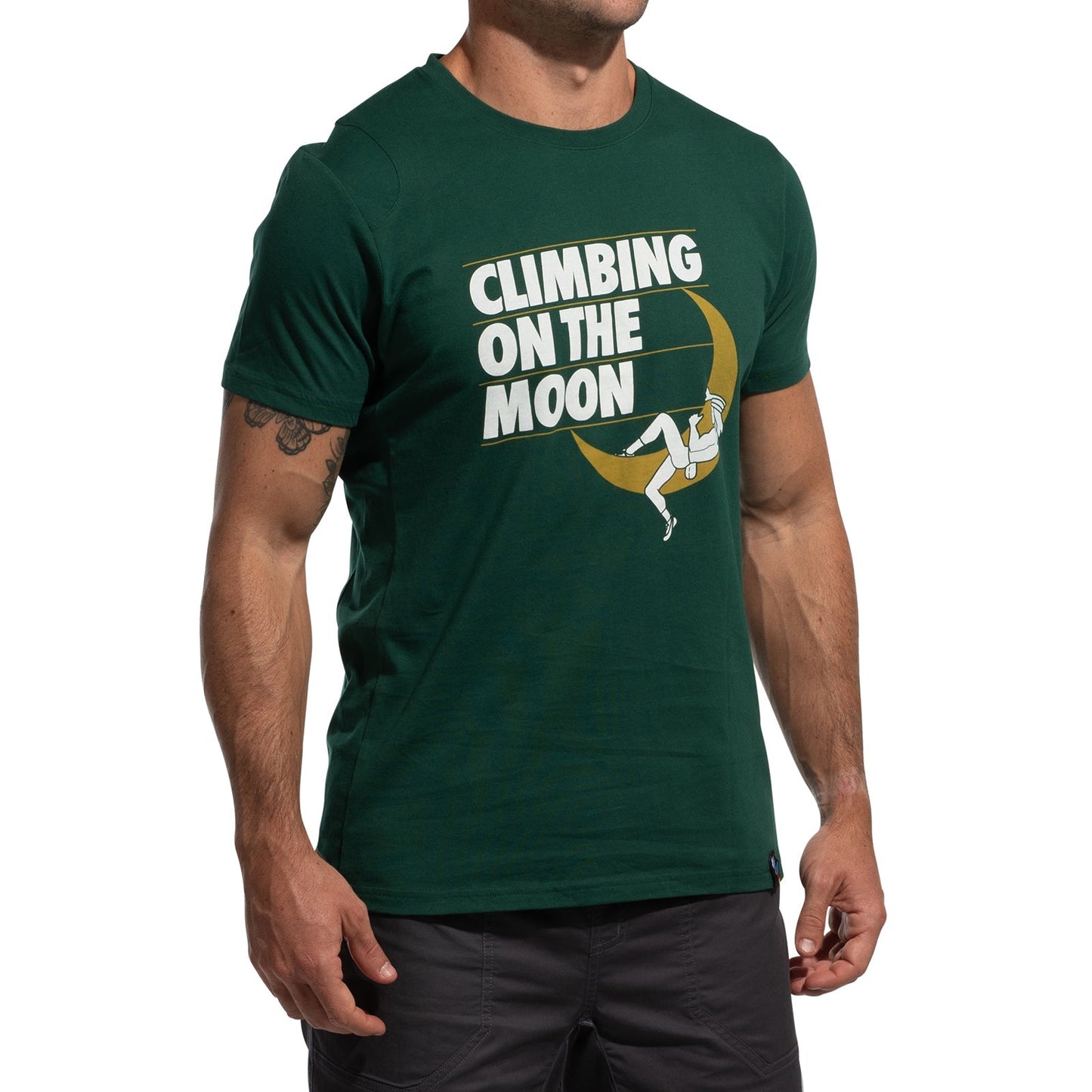 A man stands relaxed in a green La Sportiva Moon Rock T-Shirt Men's (2025), featuring "CLIMBING ON THE MOON" and a climber scaling a crescent moon.