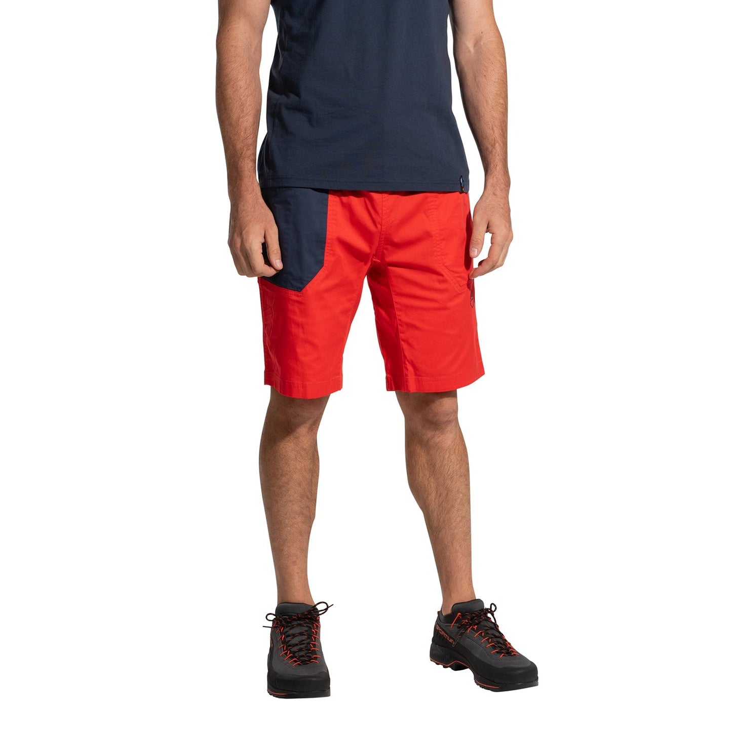 A man, head not visible, wears La Sportiva Bolt Shorts Men's (2025) in navy organic cotton with a bright red pocket and chalk bag loop, paired with black hiking shoes with red accents, standing against a white background.