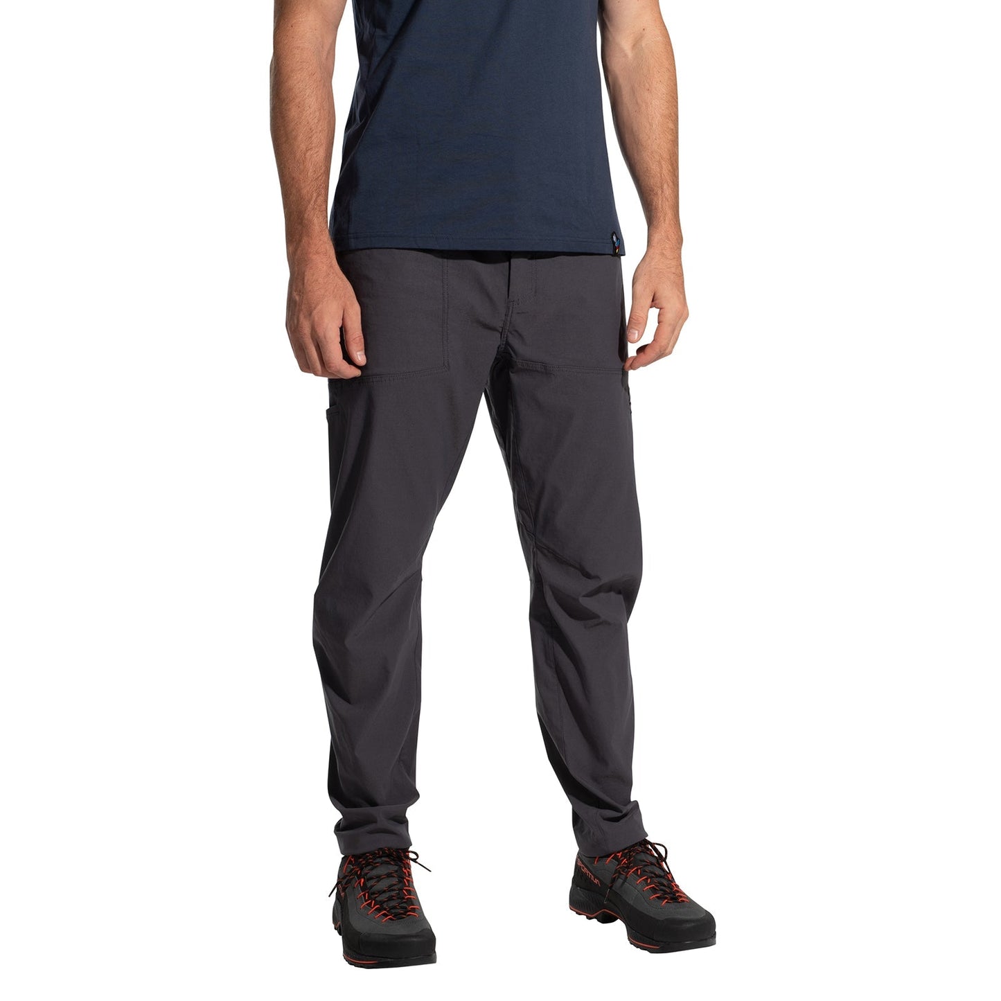 A person stands against a white background wearing a navy t-shirt, La Sportiva Talus Pants Men's (2025) in eco-friendly dark gray with articulated knees, and black hiking shoes with red laces; their face is not visible.