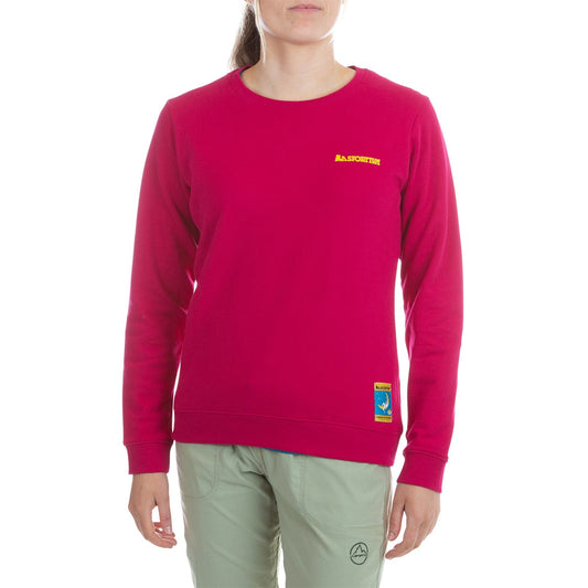 A person wearing a bright pink La Sportiva Climbing On The Moon Sweatshirt Women's (2025) with yellow logos and light green natural fiber pants stands against a plain white background, their face partially out of frame.