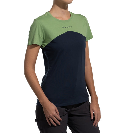 A person wears the La Sportiva Roof T-Shirt Women's (2025), featuring a light green top and dark blue bottom, paired with dark pants, as they stand against a white background.