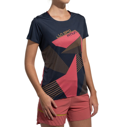 A woman wears the La Sportiva Comp T-Shirt Women's (2025), a lightweight, quick-dry climbing top with short sleeves in a navy and pink geometric pattern, paired with matching pink shorts. She stands against a white background, head partially out of frame.