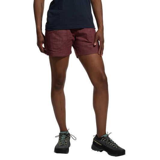 A person stands against a white background wearing La Sportiva's Sierra Rock Shorts Women's (2025) in maroon with a navy shirt and black hiking shoes featuring light blue laces, their relaxed stance highlighting comfort and ease.