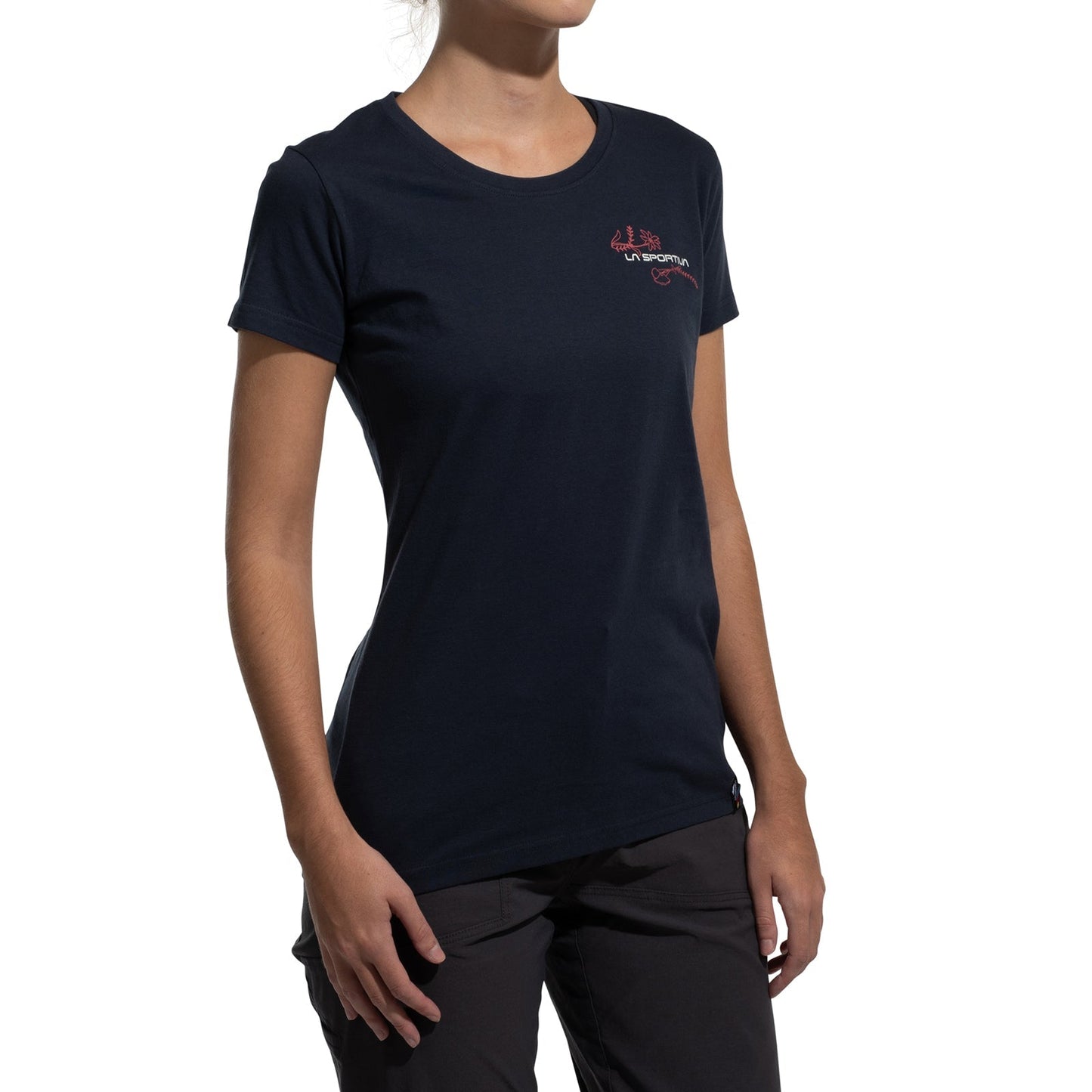A woman stands against a plain white background wearing the La Sportiva Climb Flower T-Shirt Women's (2025), a fitted short-sleeve black tee with a small red logo on the upper left chest, made from soft organic cotton.