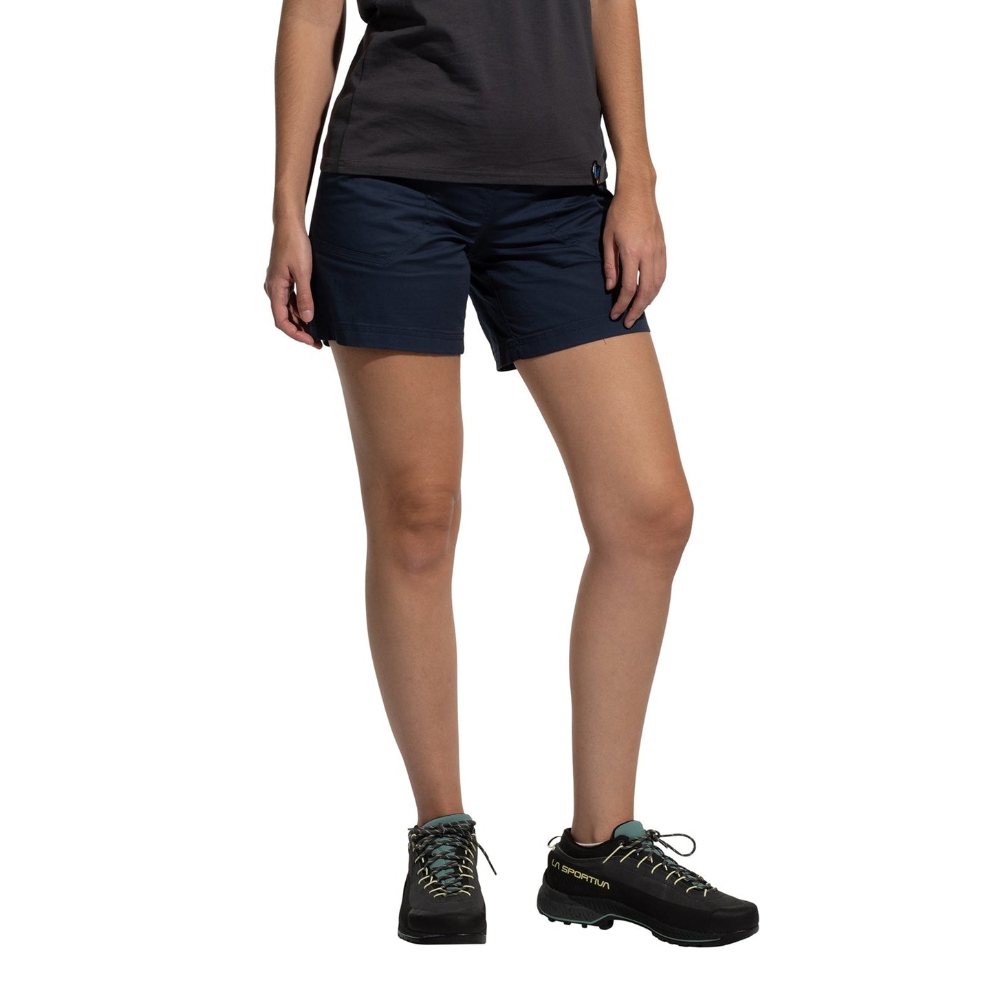 A person wearing a dark t-shirt, La Sportiva Roots Shorts Women's (2025) in navy blue with a comfortable waistband, and black hiking shoes stands against a white background, arms relaxed by their sides.