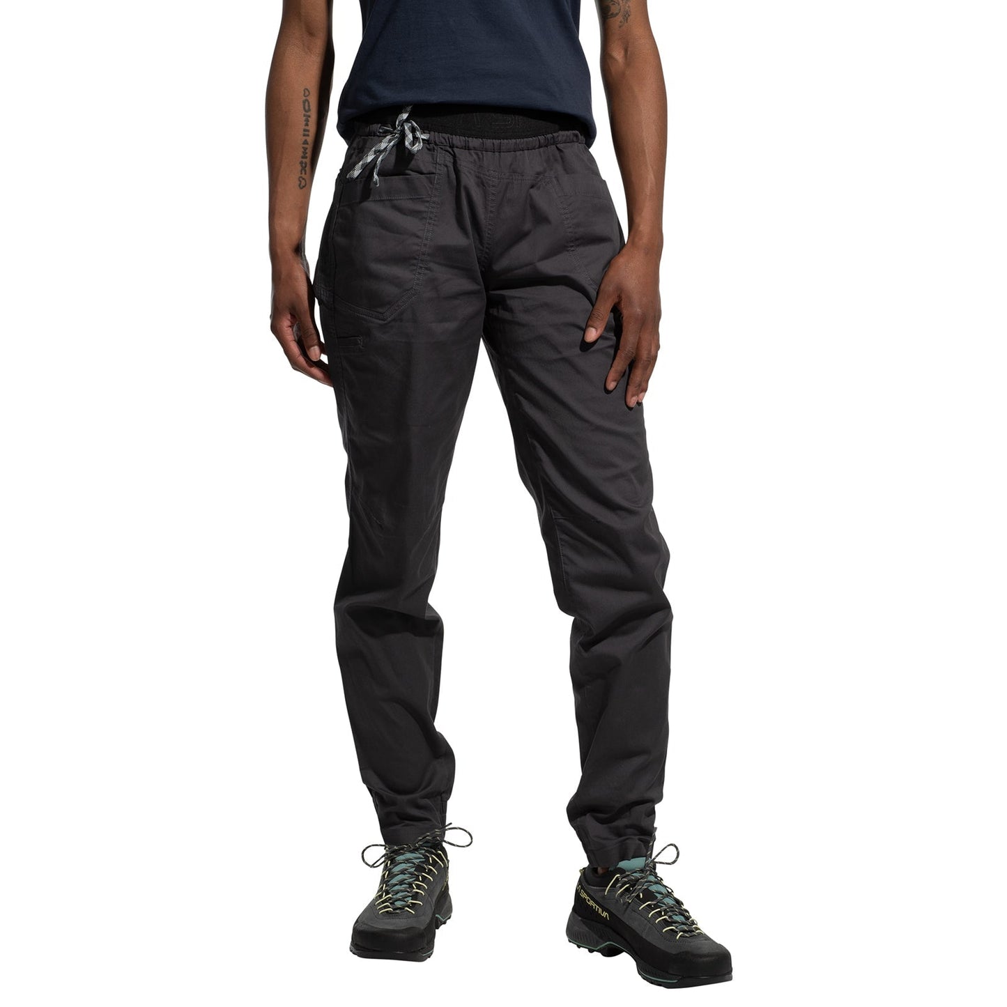 A woman stands against a white background wearing La Sportiva Tundra Pants Women's in dark gray, paired with a black T-shirt and black hiking shoes with colorful laces; her left hand rests on her thigh.