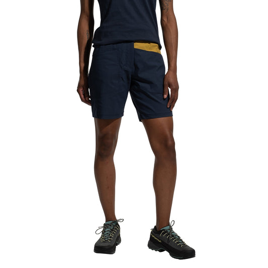 A person stands against a white background, wearing La Sportiva Mantra Shorts Women's (2025) in navy blue made from recycled fabric, paired with a matching t-shirt and black hiking shoes with light-colored laces.