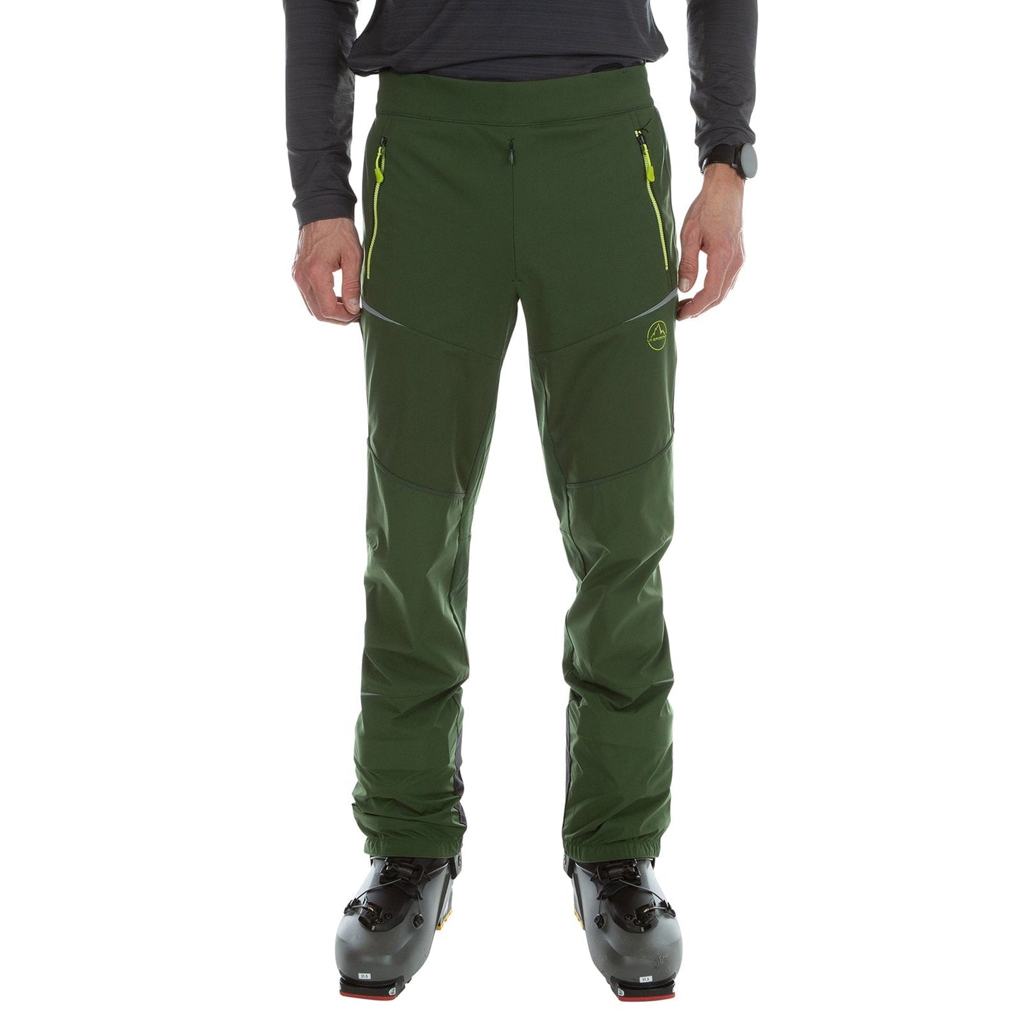 A person wearing La Sportiva Ikarus Pants Men's (2023) in dark green with yellow zipper pockets, a black long-sleeve shirt, a wristwatch, and black ski boots stands against a white background.
