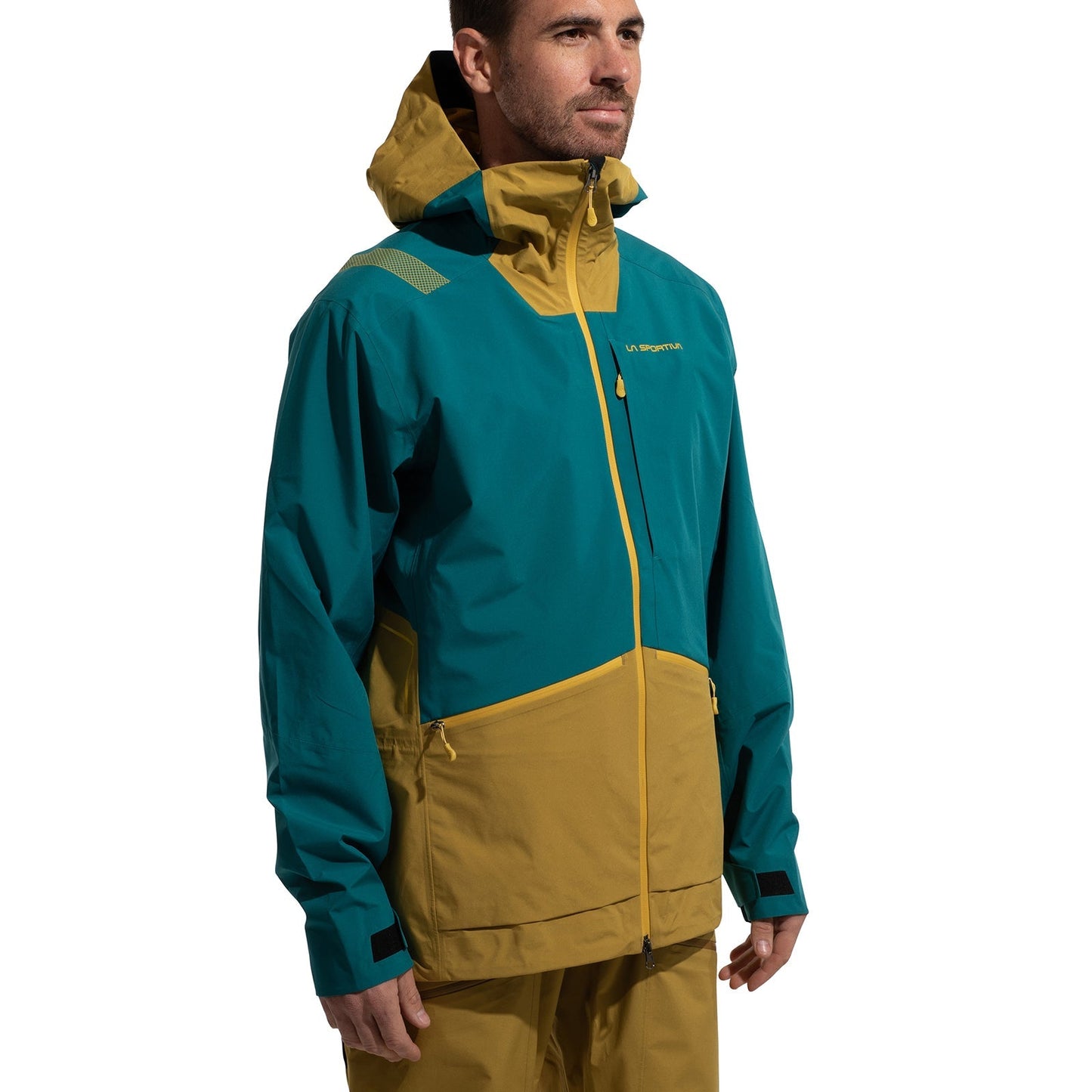 A man wears the La Sportiva Chaser EVO Shell Jacket Men’s in teal and mustard yellow with underarm ventilation, paired with matching pants. He stands slightly to the side, looking ahead with a neutral expression.