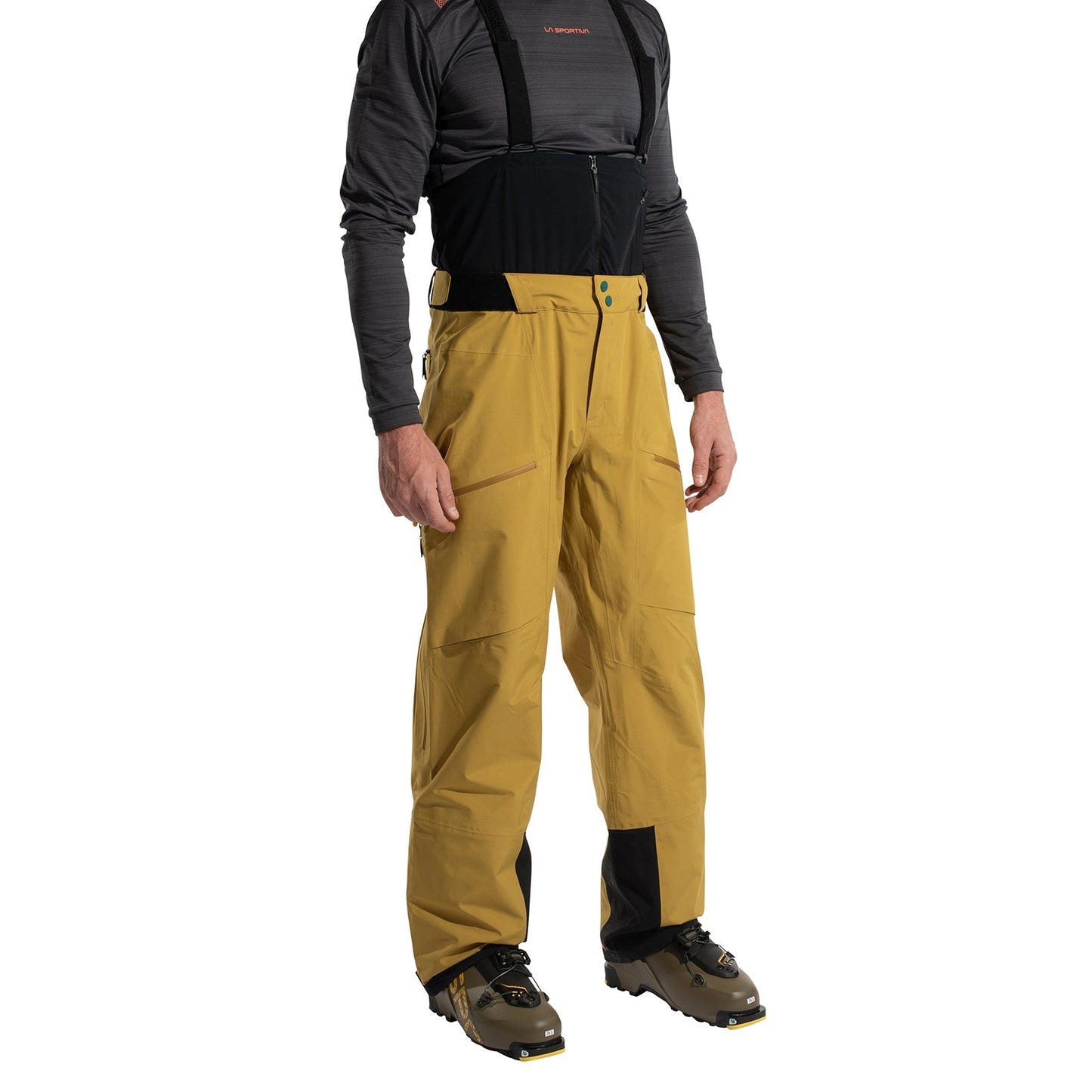 A person wearing La Sportiva Chaser EVO Shell Bib Pants Men's in mustard yellow with black suspenders, high waist, and black knee/ankle patches stands in ski boots and a dark long-sleeve shirt against a white background.