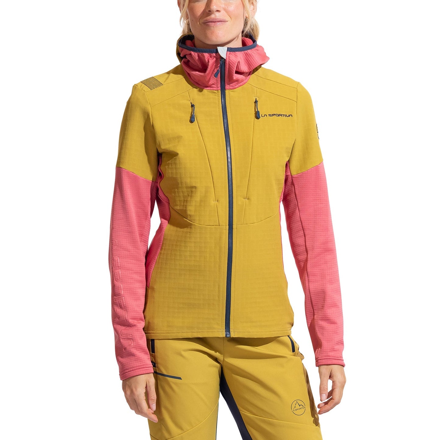 A woman in a yellow and pink La Sportiva Session Tech Hoody Women's, featuring zippered pockets, logo details, windproof protection, and a women’s-specific fit, stands facing forward in matching yellow pants.