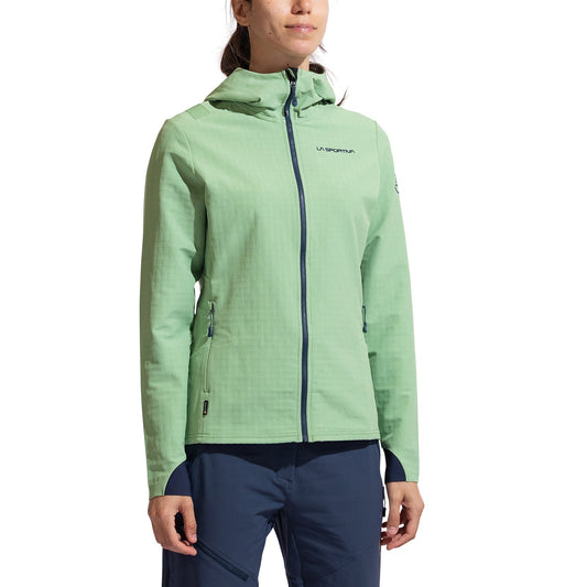 A woman stands against a plain white background wearing the La Sportiva Descender Storm Jacket Women's (2025) in light green, featuring a fleece interior, zippered pockets, and brand logos on the chest and sleeve, paired with navy blue pants.