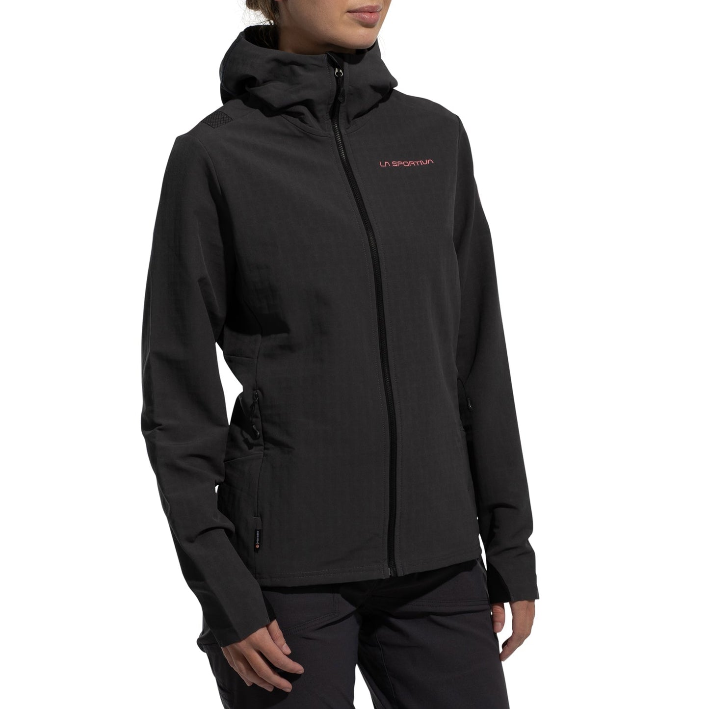 A person wearing the La Sportiva Descender Storm Jacket Women's (2025), a black softshell with fleece interior, side pockets, and front zipper, stands against a white background. The La Sportiva logo is on the chest; their face is partly out of frame.