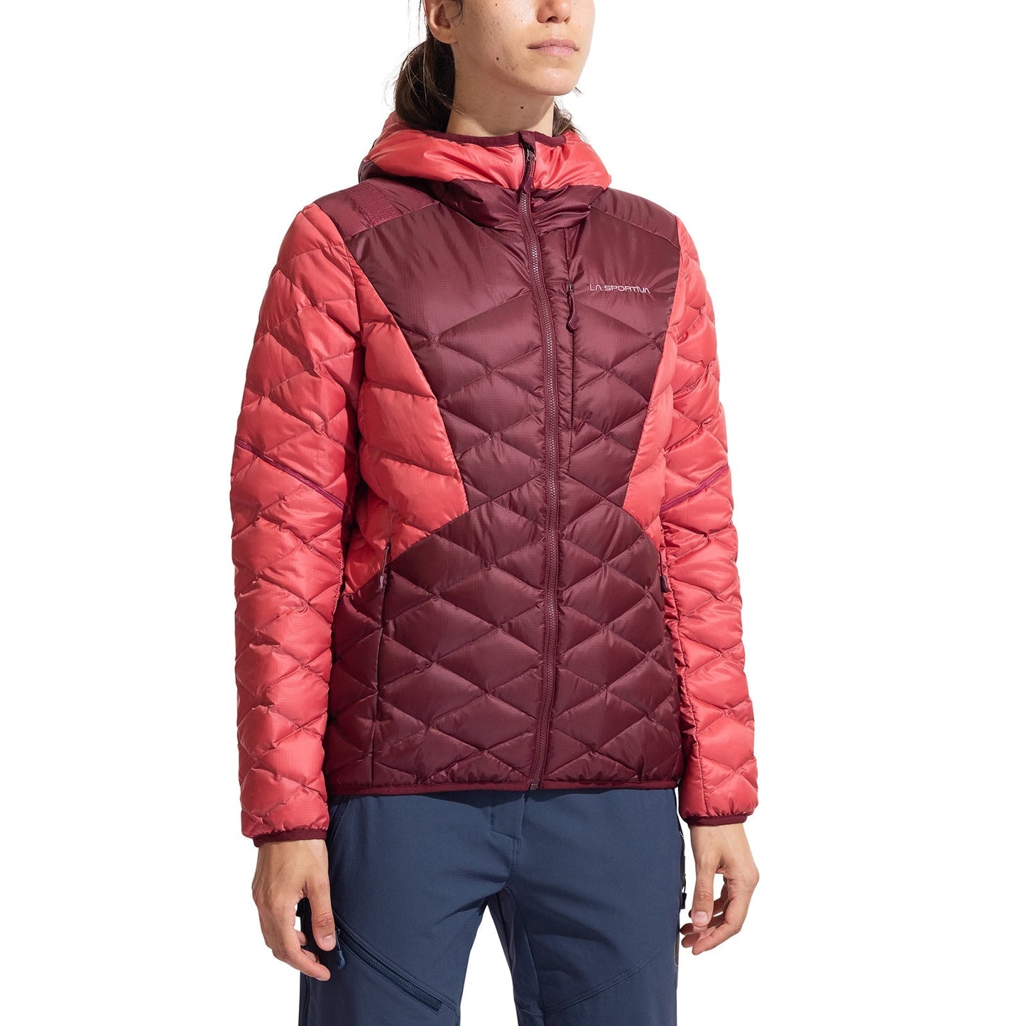 A woman wearing the La Sportiva Pinnacle Down Jacket Women's in maroon and coral with an adjustable hood and navy blue pants stands against a plain white background.
