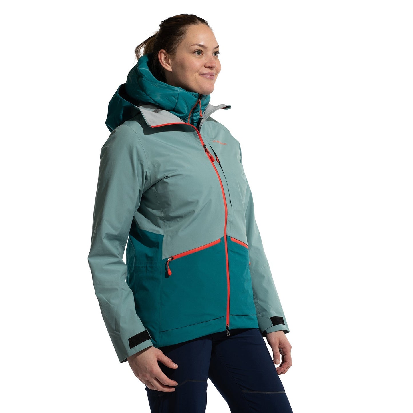 A woman wearing the La Sportiva Chaser EVO Shell Jacket Women's in two-tone teal and light blue with red zippers and reflective details stands smiling against a plain white background.