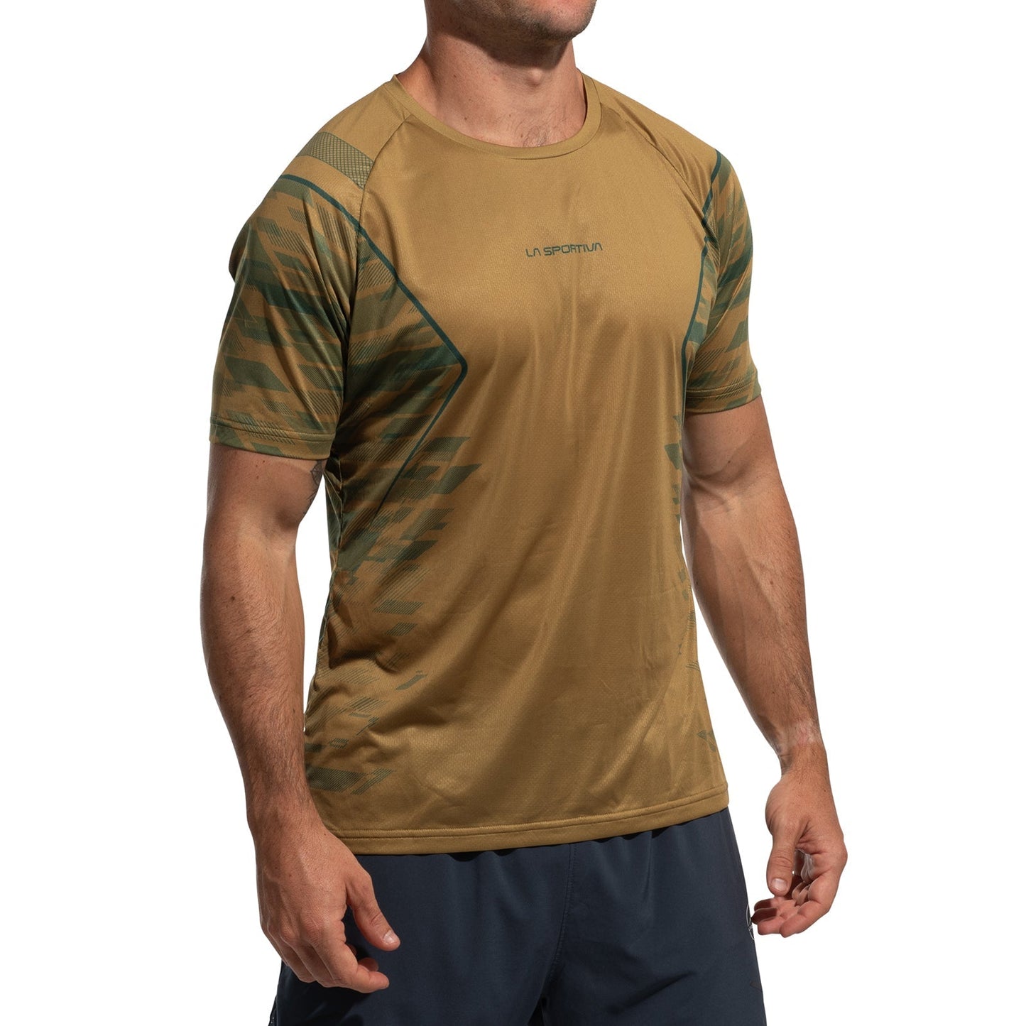 A man stands against a white background wearing the La Sportiva Pacer T-Shirt Men's—a short-sleeved, light brown ultralight top with green patterns and the La Sportiva logo on the chest—paired with dark shorts.