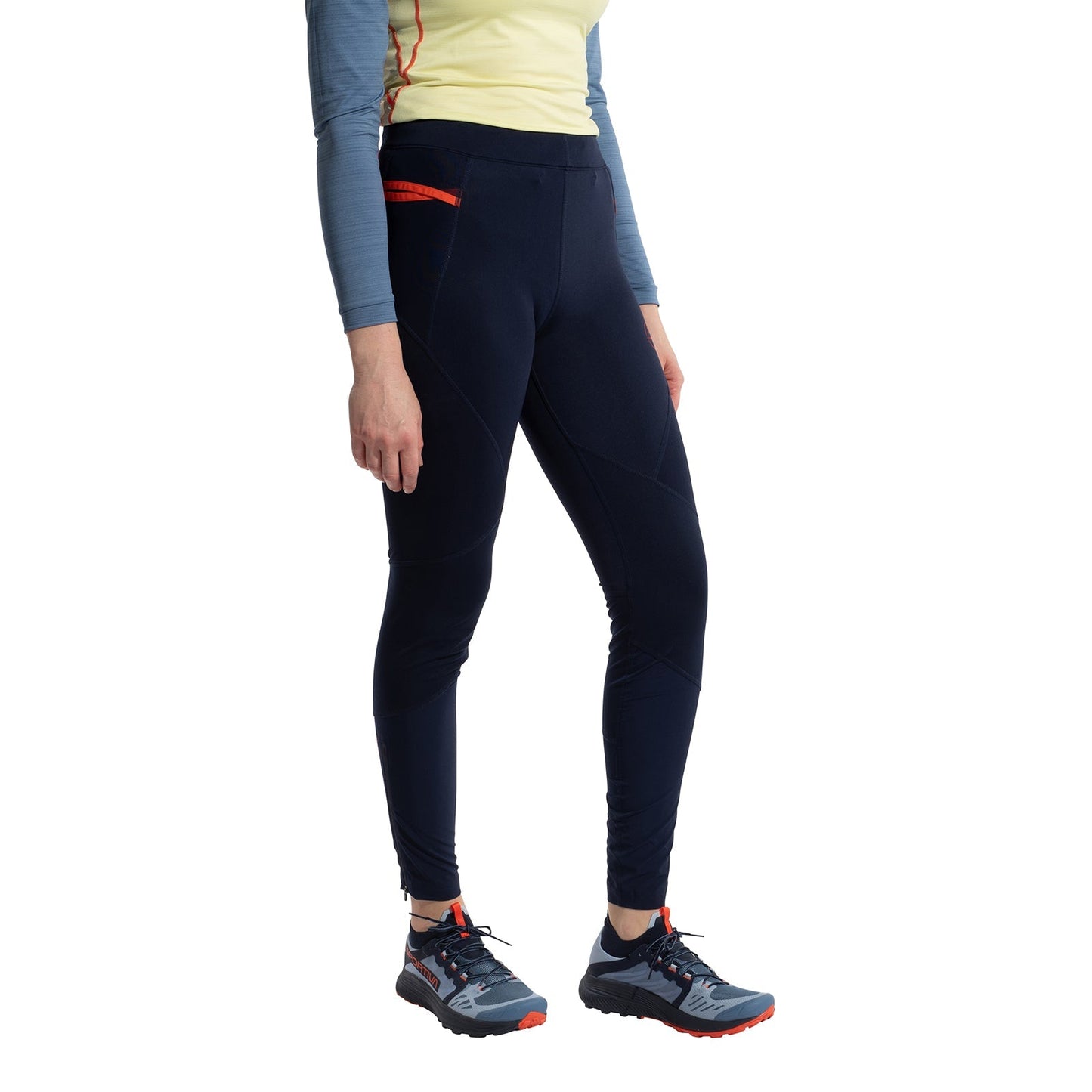 A woman wearing a light yellow long-sleeve top, La Sportiva Primal Pants Women's in dark navy with red accents for winter running, and gray trail running shoes with orange details, stands against a white background.