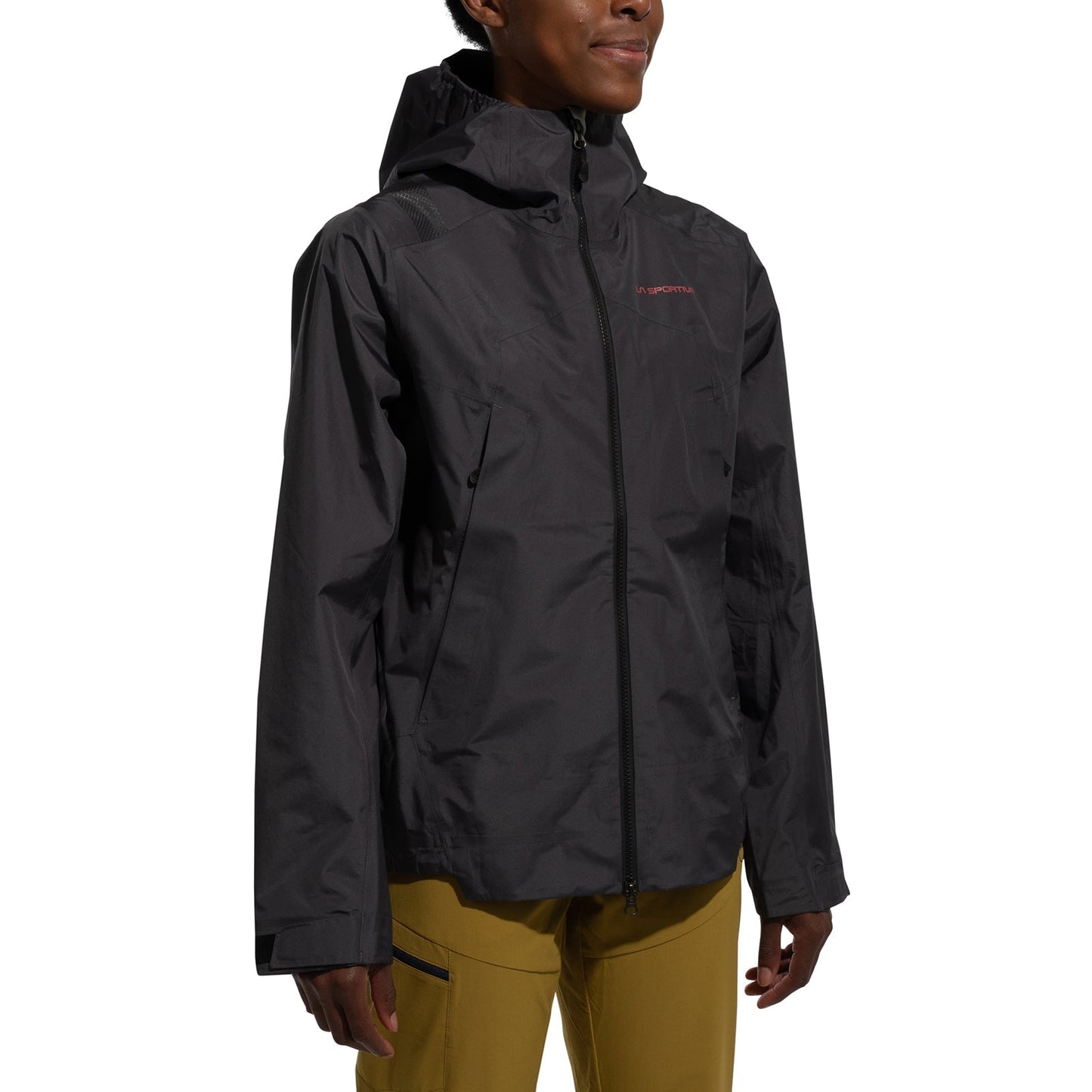 A woman wearing the La Sportiva Discover Shell Jacket Women's (2025) in dark gray, featuring an adjustable hood and front zipper, paired with mustard yellow pants, stands against a plain white background.