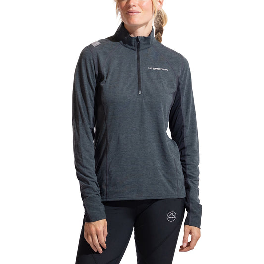 A person stands against a white background wearing La Sportiva’s Swift Long Sleeve Women's shirt in dark gray and black pants with visible brand logos—an ideal winter Mountain Running outfit.