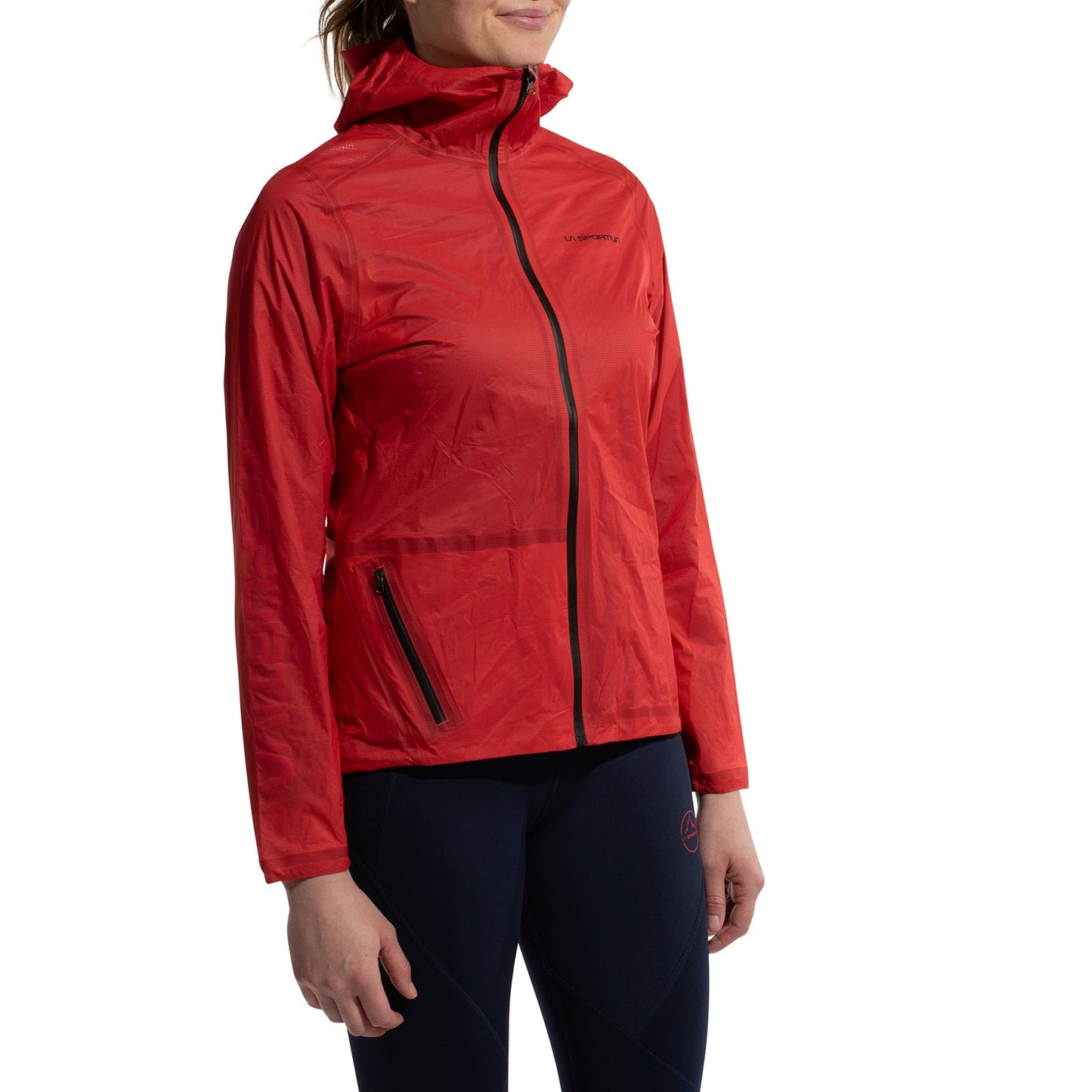 A woman models the La Sportiva Ethereal Pro Jacket Women's—a bright red, rainproof shell with a hood and zippered pockets—over dark navy leggings, standing against a plain white background, ready for any weather.