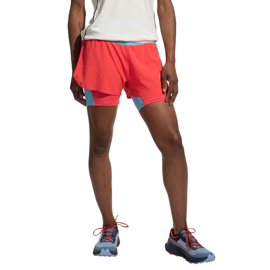 A woman wears La Sportiva Ultra Distance Shorts in bright red with blue trim, paired with a white shirt and blue-red running shoes, standing with one leg slightly bent against a plain white background.