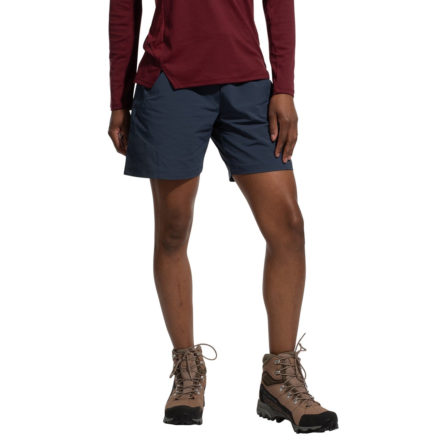A person wearing a long-sleeve maroon shirt, La Sportiva Trail Guard Shorts Women's (2025), and tan hiking boots stands with one hand in a pocket against a plain white background.