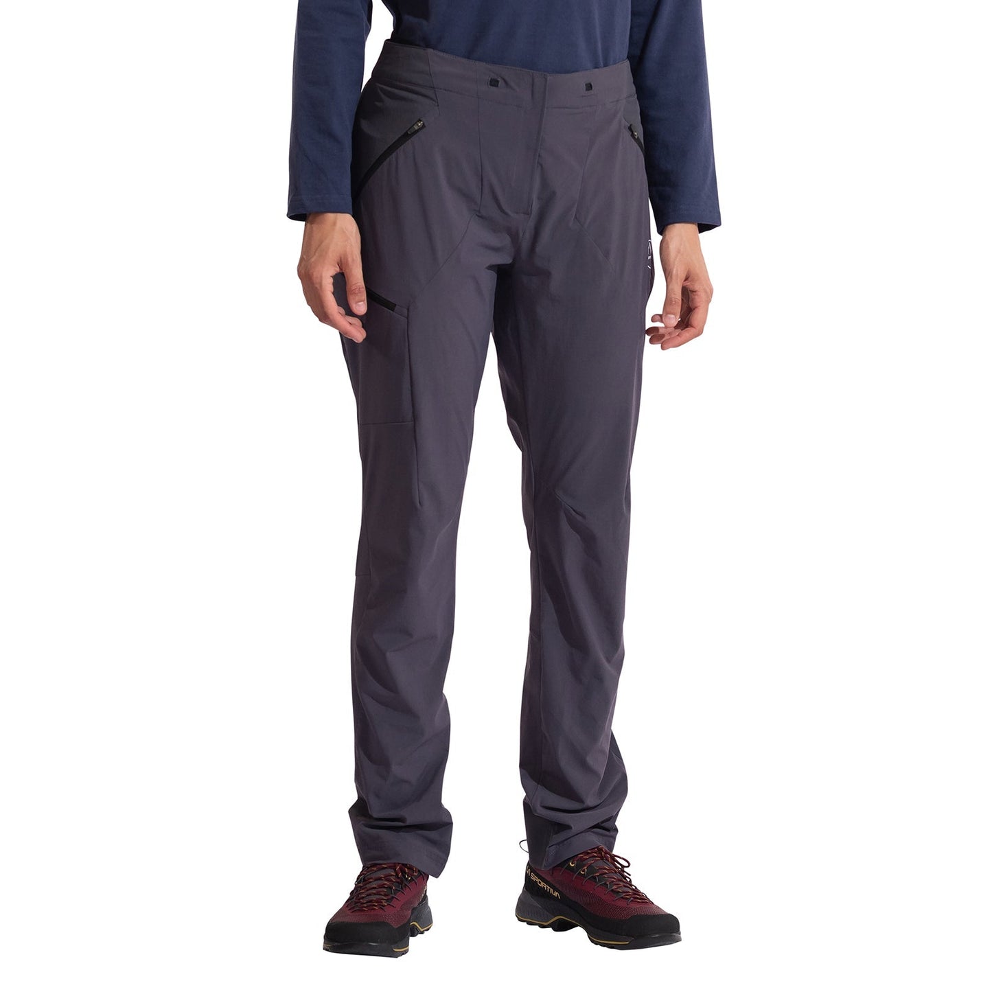 A woman stands facing forward against a white background wearing La Sportiva Monument Pants Women's (2025), which feature zippered pockets and a tailored fit, paired with a navy long-sleeve shirt and maroon hiking boots.