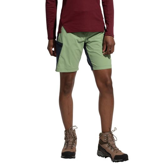 A person stands against a white background wearing a maroon long-sleeve shirt, brown hiking boots, and La Sportiva West Crest Shorts Women's (2025) in light green and dark blue. The person's head is not visible.