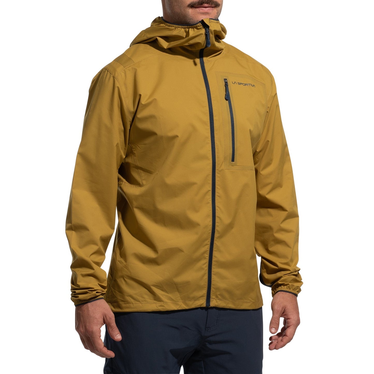 A man wearing the La Sportiva Wall Breeze Stretch Jacket Men's (2025) in mustard yellow, featuring wind protection, front zipper, chest pocket, elastic cuffs, logo on the left chest, helmet-compatible hood, and dark pants.