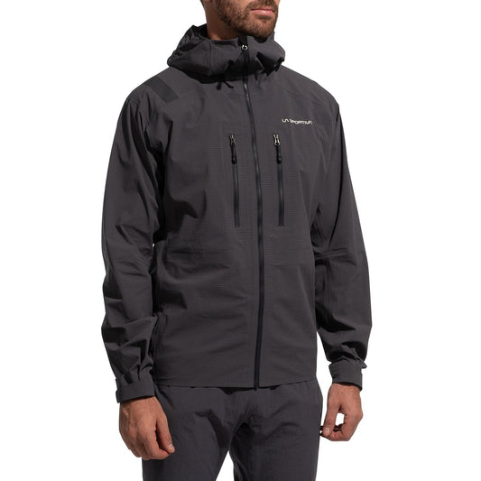 A man stands with arms relaxed, wearing the La Sportiva Helixir Shell Jacket Men's (2025) in dark gray with zippered chest pockets, matching pants, and a subtle logo on the left chest.