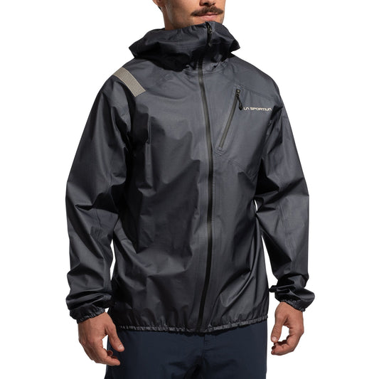 A man wearing the La Sportiva Aequilibrium Lite GTX Jacket Men's in dark gray, featuring a hood, front zipper, chest pocket, elastic cuffs, a left chest logo, and a reflective right shoulder stripe—ideal for summit adventures.