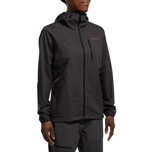A woman wearing the La Sportiva Wall Breeze Stretch Jacket in black, featuring a helmet-compatible hood, chest zipper pocket, and small logo on the left side, with matching pants, stands against a white background.