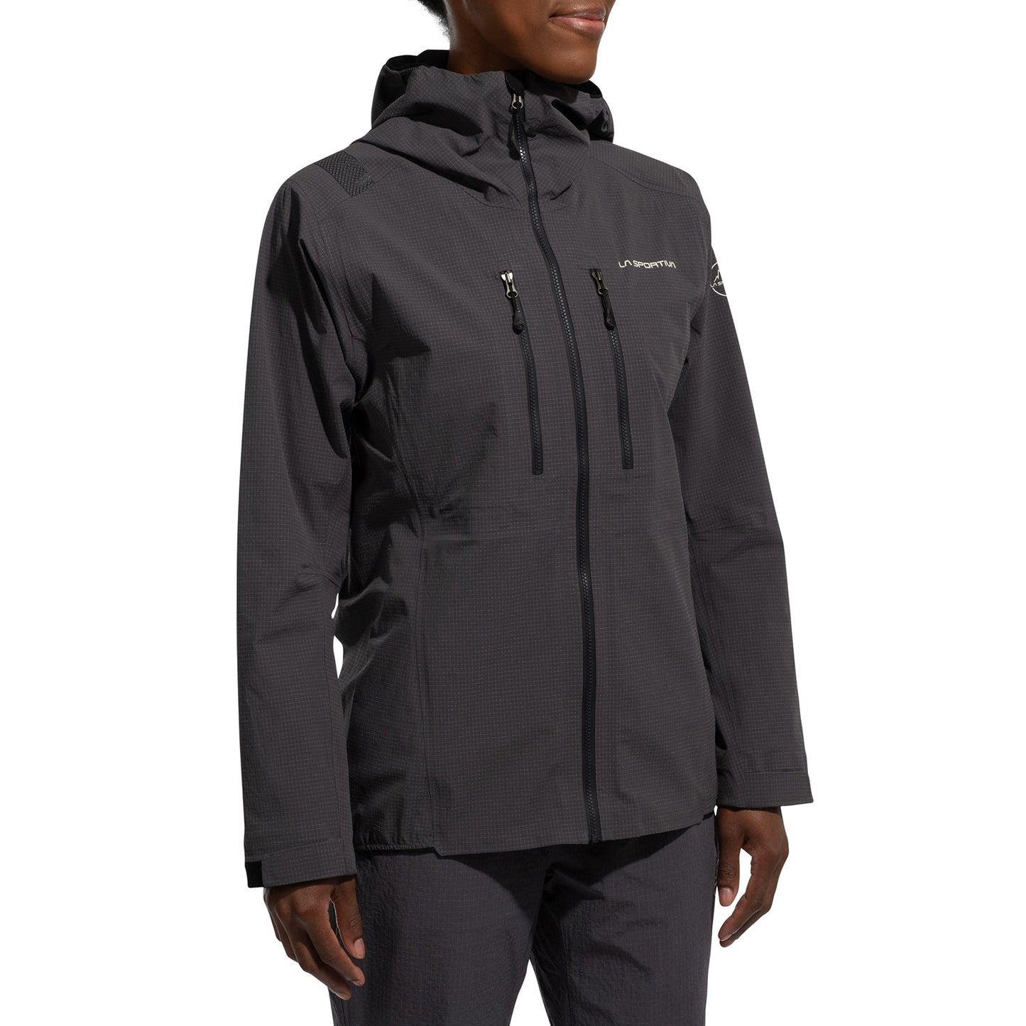 A woman stands against a plain white background wearing the dark gray La Sportiva Helixir Shell Jacket (2025), featuring a hood, waterproof Spectra fibers, and zippered chest pockets, paired with matching pants—ideal attire for big wall adventures.