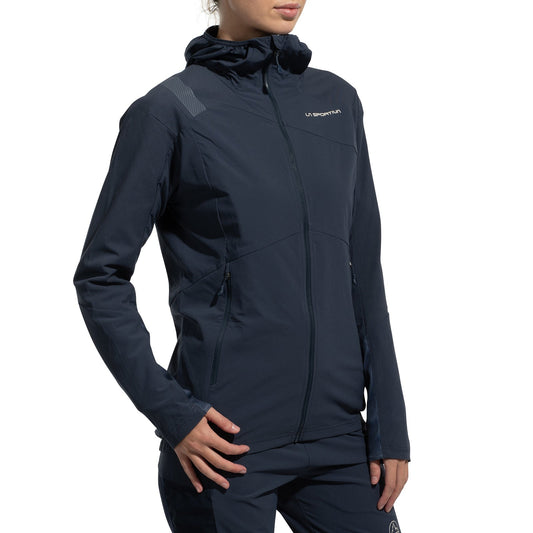A woman wears the La Sportiva Aequilibrium Speed Jacket (2025) in dark navy, featuring a hood, zip front, pockets, small chest logo, and subtle shoulder design. The breathable jacket is paired with matching pants against a white background.