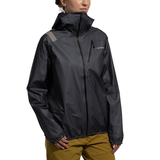 A person wears the La Sportiva Aequilibrium Lite GTX Jacket Women's in dark gray, featuring a front zipper, small chest logo, and striped shoulder accent, paired with light brown pants against a plain white background.