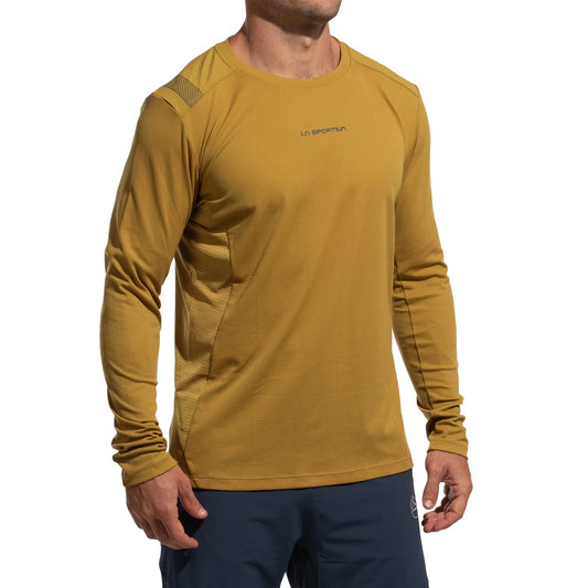 A person wears a mustard yellow La Sportiva Beyond Long Sleeve Men's (2025) top with lightweight Polartec Delta Cooling Fabric and navy pants, standing with arms relaxed against a white background. The head is partially out of frame.