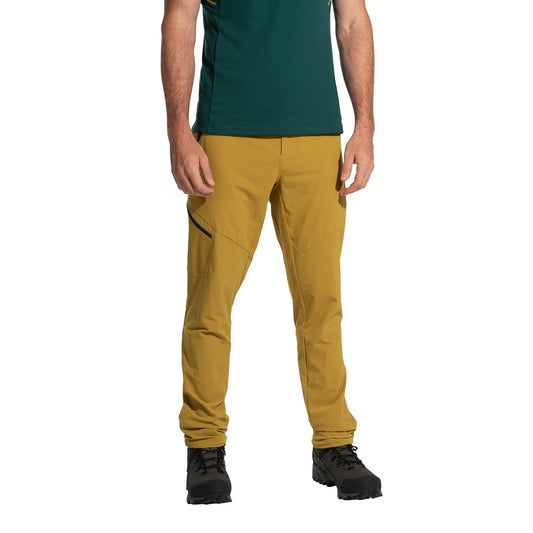 A person wears La Sportiva West Crest Pants Men's (2025) in mustard yellow with zippered pockets, paired with a green short-sleeve shirt and black hiking boots, standing against a white background. The photo is cropped at the shoulders.