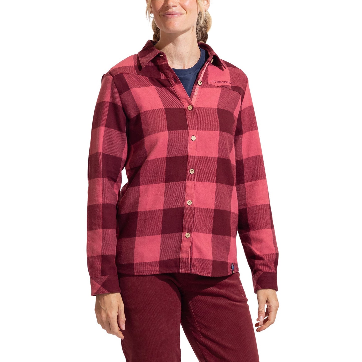 A woman wearing the La Sportiva Rambler Flannel Shirt in a red and pink plaid, featuring coconut buttons, is standing with her arms relaxed by her sides and smiling slightly over a dark undershirt and red pants.