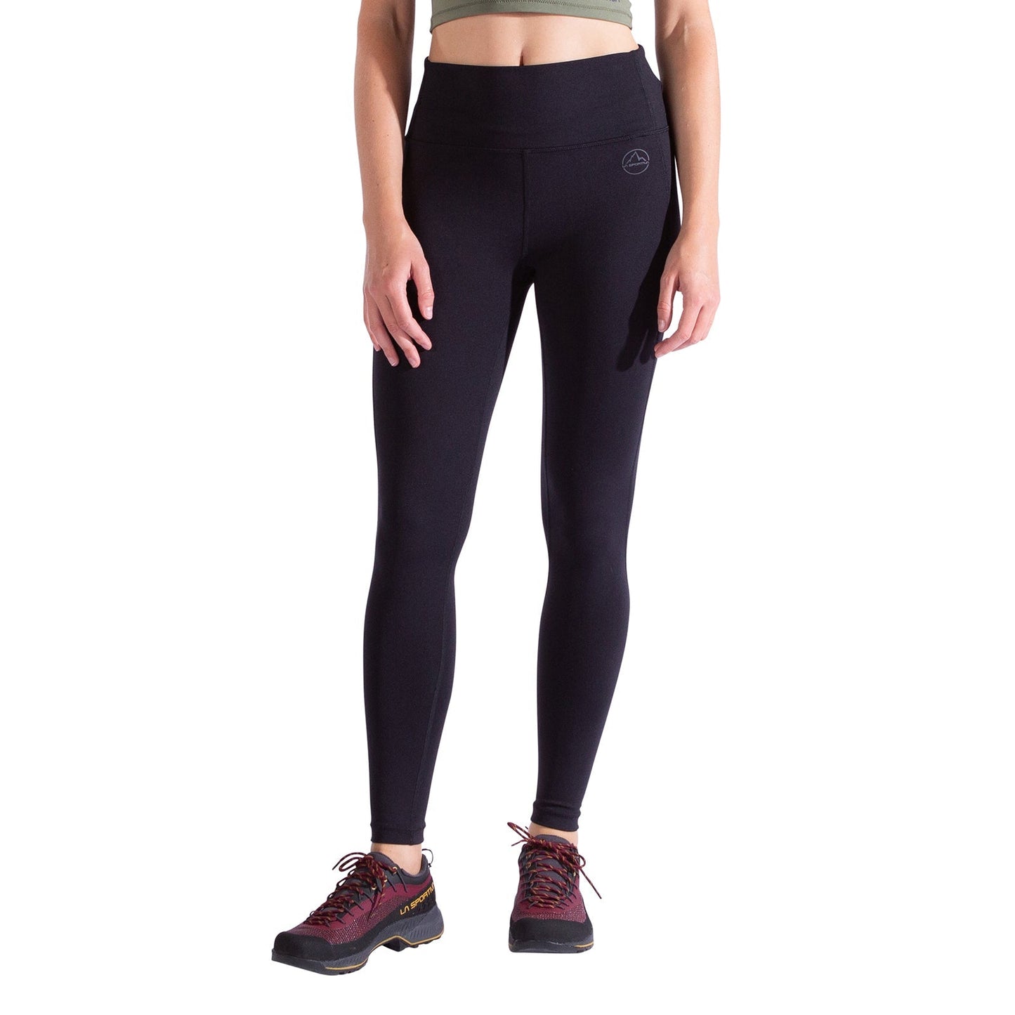 A woman stands against a white background, showcasing only her lower torso and legs in high-waisted black La Sportiva Cosmosis Leggings, paired with dark hiking shoes and a light green crop top. The image is cropped at the shoulders.
