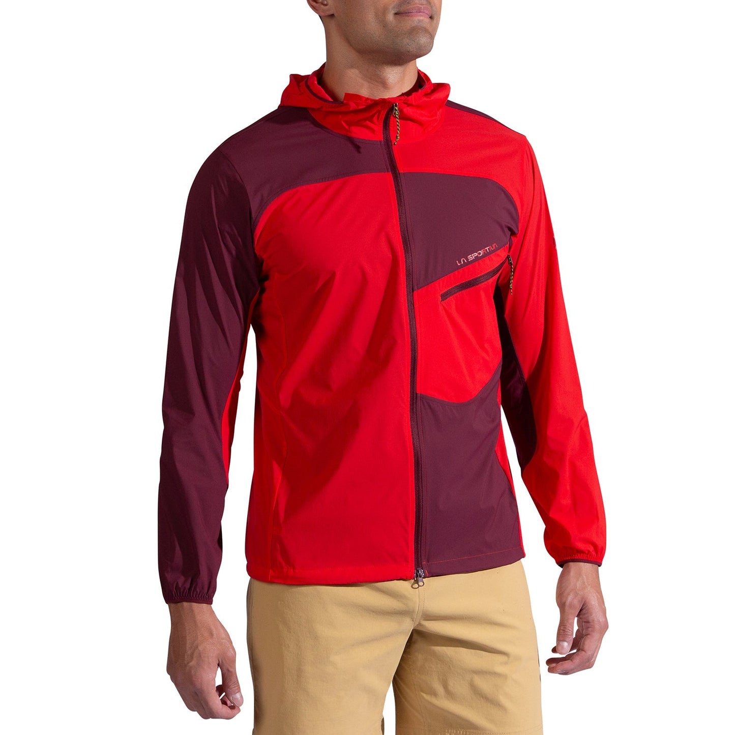 A man models the La Sportiva Freeway Wind Jacket Men's in red and burgundy, with a front zipper and chest pocket, paired with beige shorts. He stands slightly turned to the side against a white backdrop.