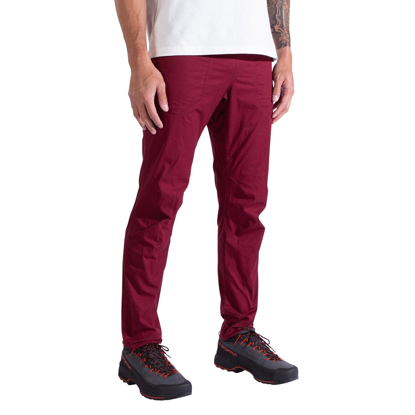 A person wearing La Sportiva Roots Pants Men's in slim-fit burgundy, a white organic cotton t-shirt, and black hiking shoes with red accents stands against a plain white background.