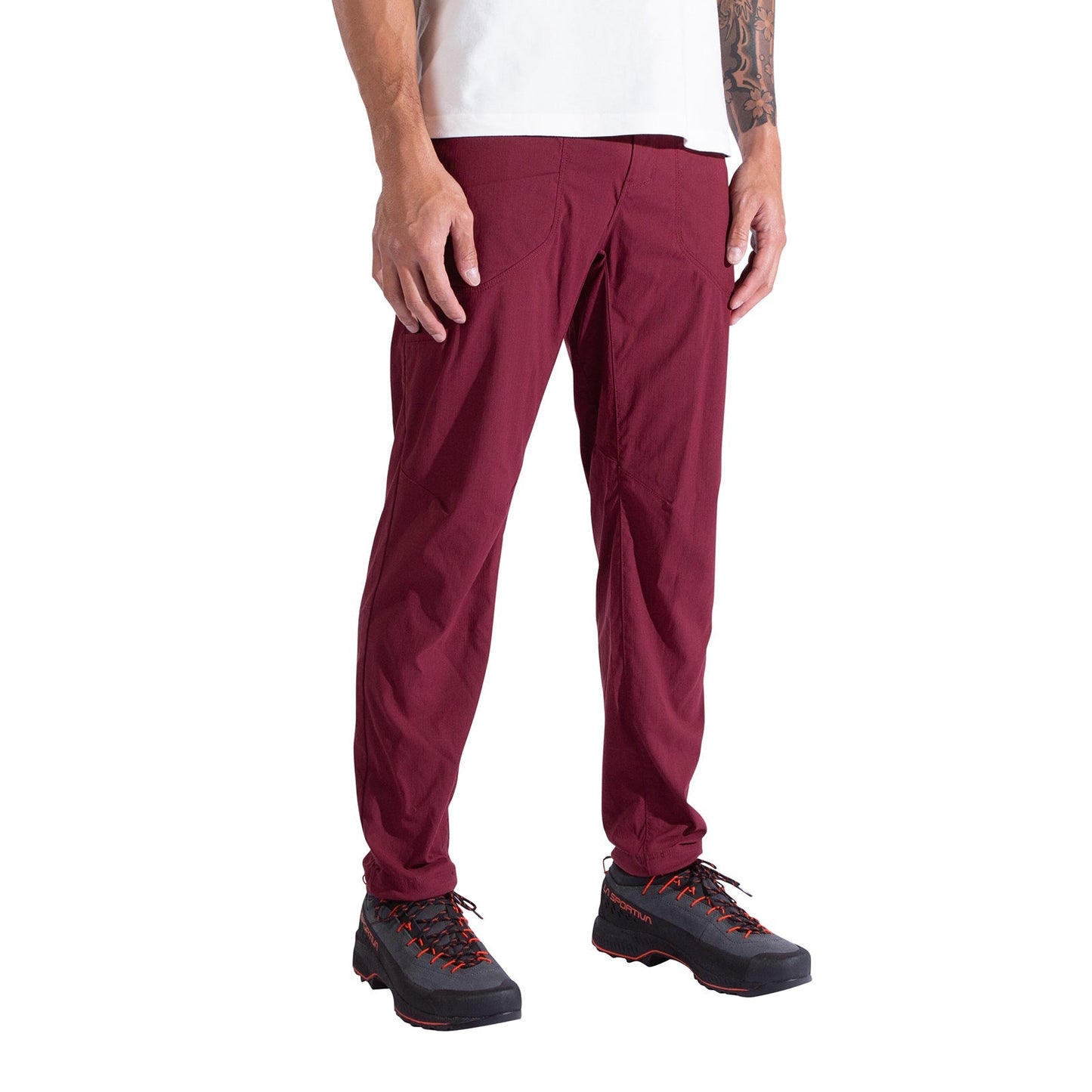 A person stands against a white background, wearing La Sportiva Labyrinth Pants Men's in burgundy with an adjustable waistband, paired with a white shirt and black hiking shoes featuring orange laces. Their arms rest naturally at their sides.