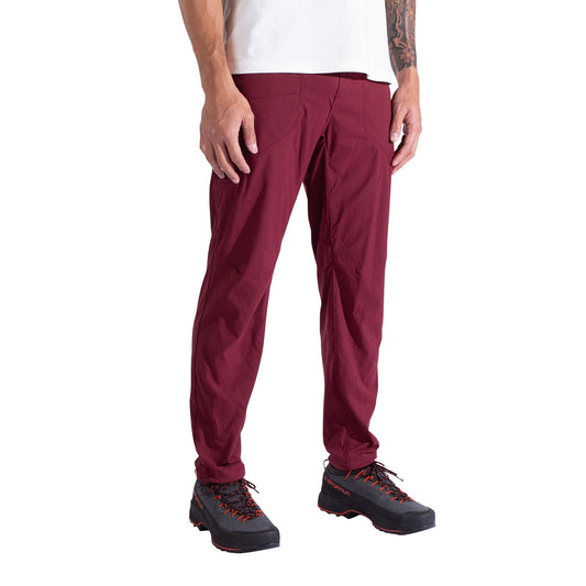 A person stands against a white background, wearing La Sportiva Labyrinth Pants Men's in burgundy with an adjustable waistband, paired with a white shirt and black hiking shoes featuring orange laces. Their arms rest naturally at their sides.