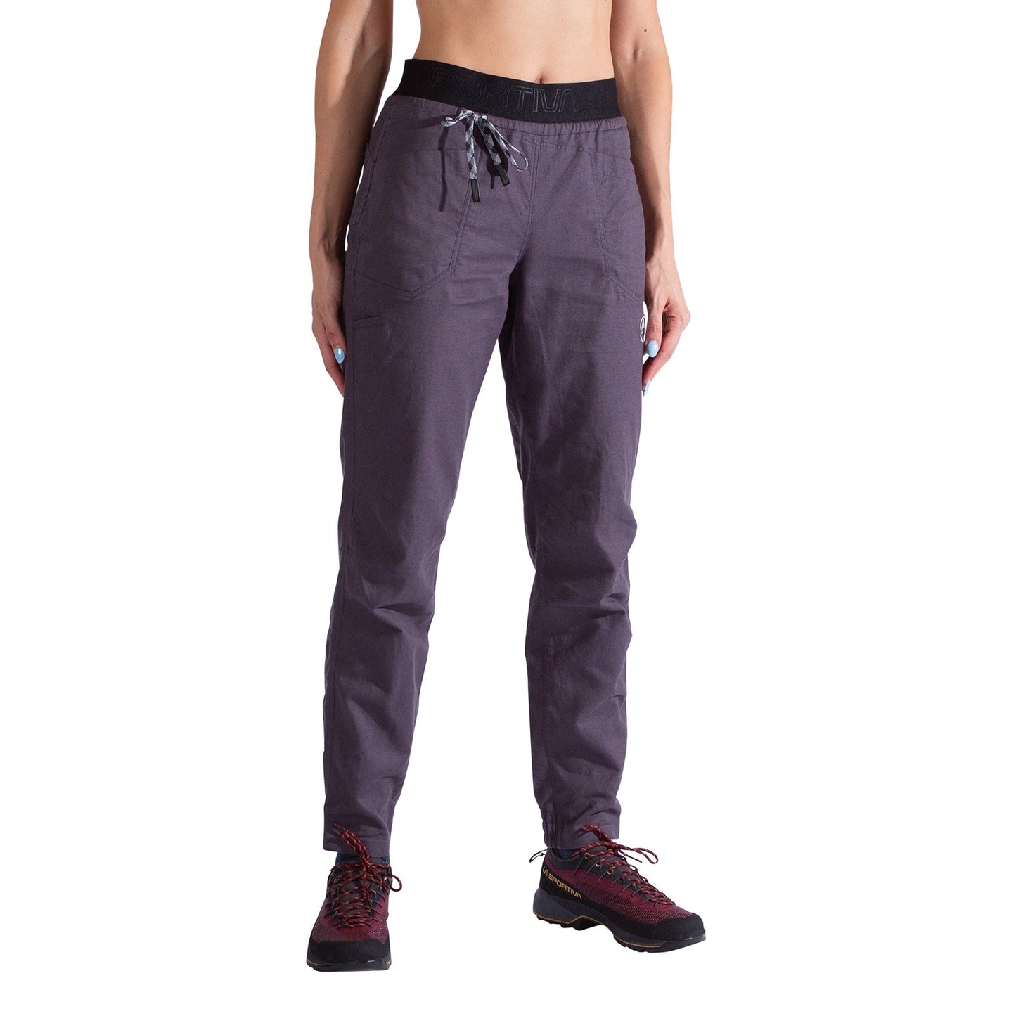 A person wears La Sportiva Bolt Pants Women's in purple organic cotton with an adjustable black waistband, drawstring, and pockets, paired with maroon hiking shoes, standing against a white background.