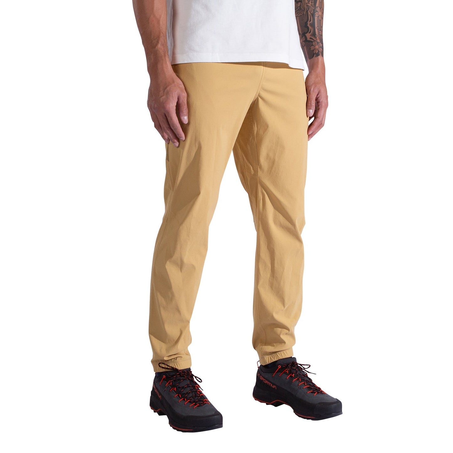 A person wears La Sportiva Gambit Pants Men's in tan, a white T-shirt, and black hiking shoes with red accents, standing against a plain white background. The person's head is not visible in the image.