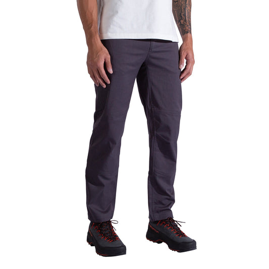 A person stands against a white background, showing only the lower half of their body and wearing La Sportiva Mainliner Pants Men's in dark gray, along with a white t-shirt and black hiking boots with red laces.